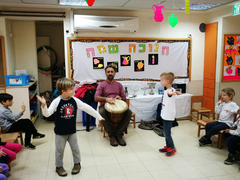 ילדים בגן רוקדים לצלילי תוף ג׳מבה מול שלט חנוכה שמח
